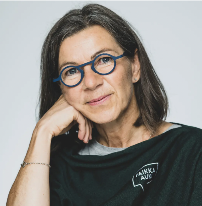 Jaana Toikkanen hymyilee kameralle ja nojaa käteensä.